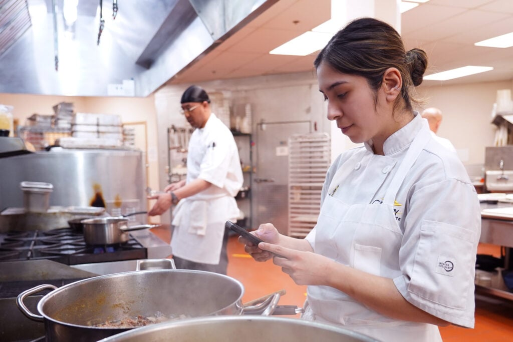 A culinary student looks at a phone while standing next to large cooking pots in a commercial kitchen with another student working in the background