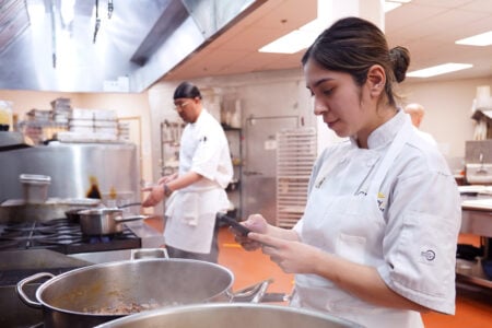 A culinary student looks at a phone while standing next to large cooking pots in a commercial kitchen with another student working in the background