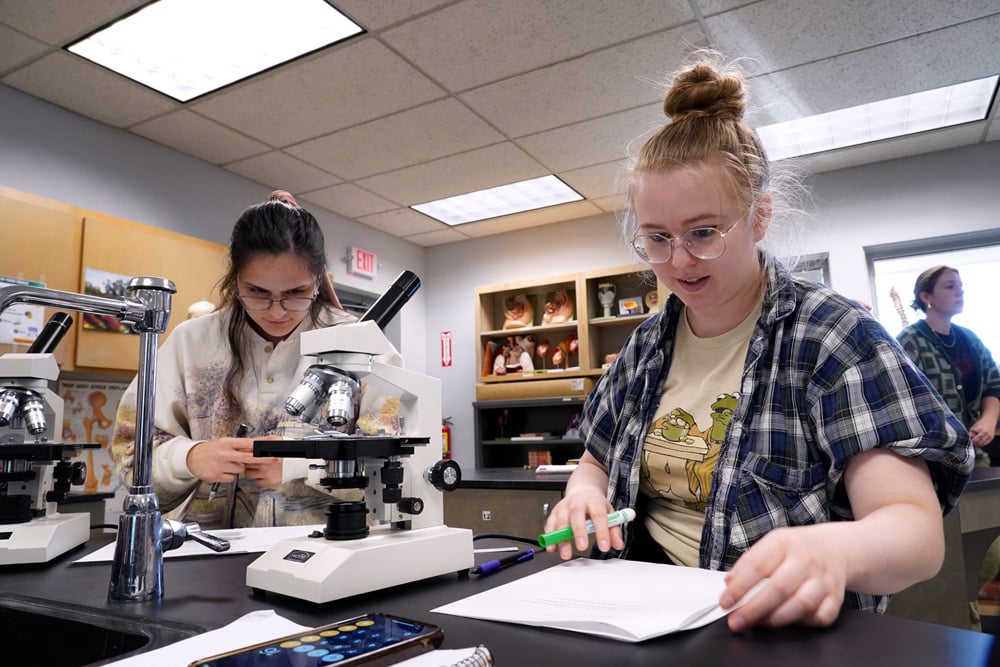 Two students working with microscopes in a science laboratory, with another person in the background.