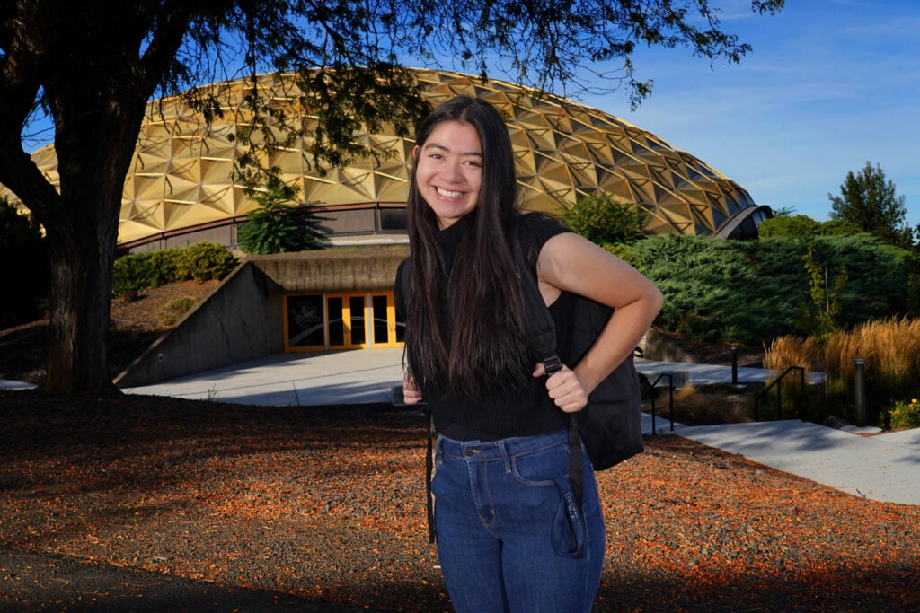 Young woman with a backpack smiling in front of a domed campus building on a sunny day.