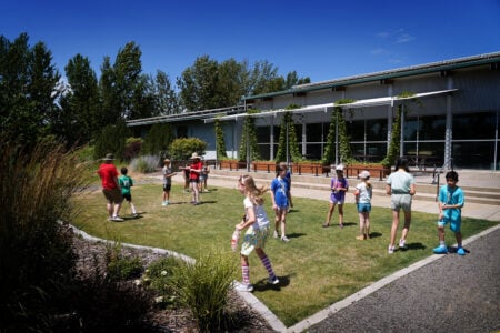 Children participate in an outdoor Kids College activity on a grassy lawn beside a modern educational building. The group gathers in small clusters while instructors supervise, with landscaped gardens, trees, and a clear blue sky surrounding the campus setting.