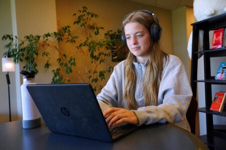 Person wearing over-ear headphones using an HP laptop at a table with a water bottle, indoors with plants and bookshelves in the background.