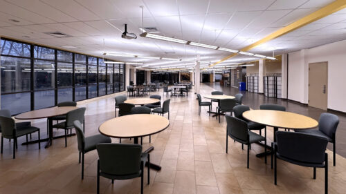 A spacious open dining area with round tables and padded gray chairs on tan tile flooring. Floor-to-ceiling windows line the left wall. Ceiling-mounted projector visible. Gold accent trim on ceiling. WWCC colors (black and gold) throughout.