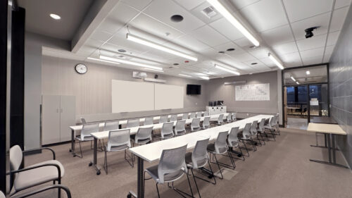 A modern classroom with long white tables and light gray mesh chairs arranged in rows. Dual whiteboards and a wall-mounted TV at the front. A periodic table poster is displayed on the right wall. Gray carpeted floor with a glass partition on the right side.