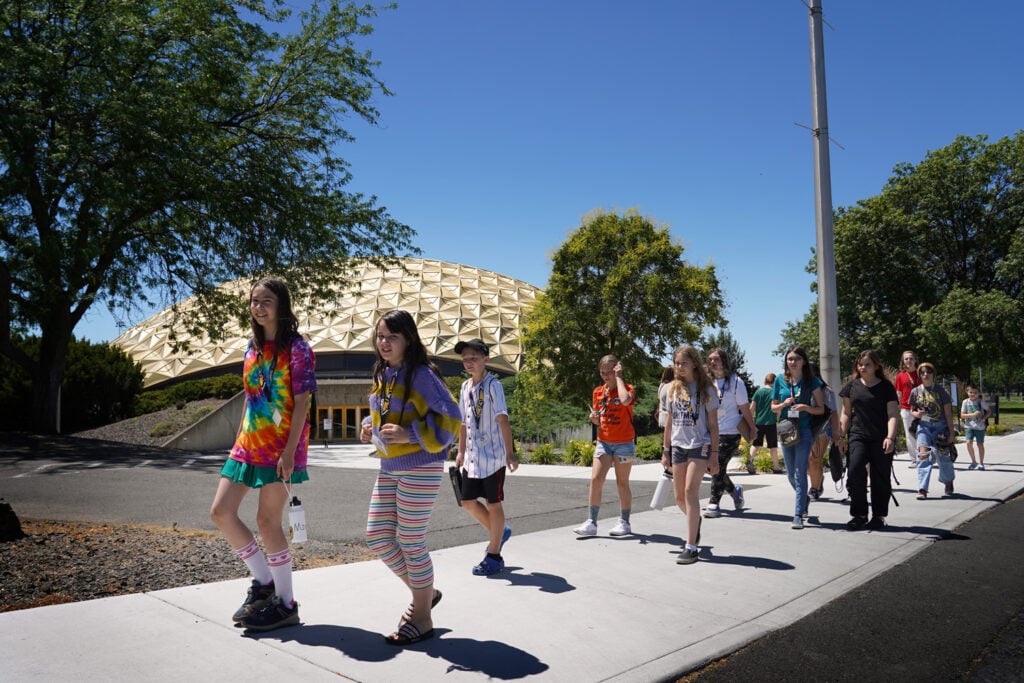 A group of Kids College participants walks along a campus sidewalk during an outdoor learning activity. Trees and landscaped grounds surround a distinctive dome-shaped building in the background as students move together across the college campus on a sunny day.