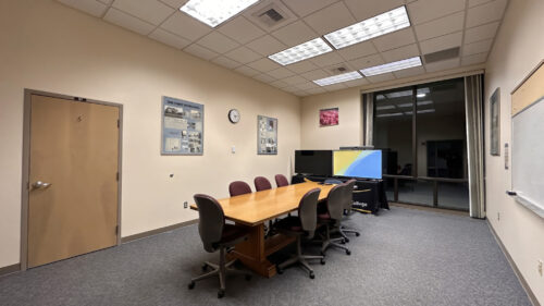 A small conference room with a rectangular wood table and six rolling chairs. A flat-screen display sits at the end of the table. Historical photo displays and artwork hang on the walls. A whiteboard is mounted on the right. Glass window looks into an adjacent space.