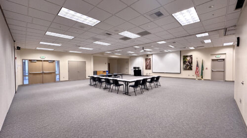 A large multipurpose room with gray carpet and high ceilings. Folding tables are arranged in a square configuration with stackable chairs. A pull-down projection screen and ceiling-mounted projector are at the front. US and Washington State flags flank the screen. Wildlife photography decorates the walls.