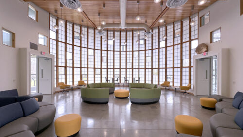A dramatic circular rotunda with soaring white walls and a full-height grid of translucent windows creating a lantern effect. Polished concrete floors. Circular green upholstered seating, yellow ottomans, and blue-gray sofas are arranged throughout. Wood-slat ceiling with pendant lights.