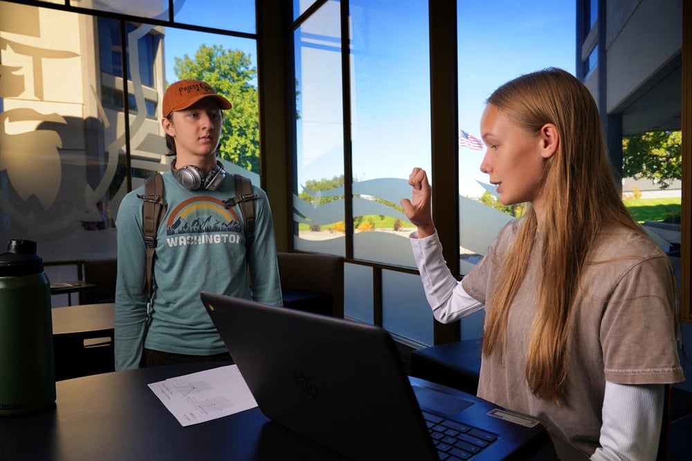 Two people at a table with a laptop in a bright room with large windows; one person gestures while speaking and the other stands nearby wearing a backpack and headphones.